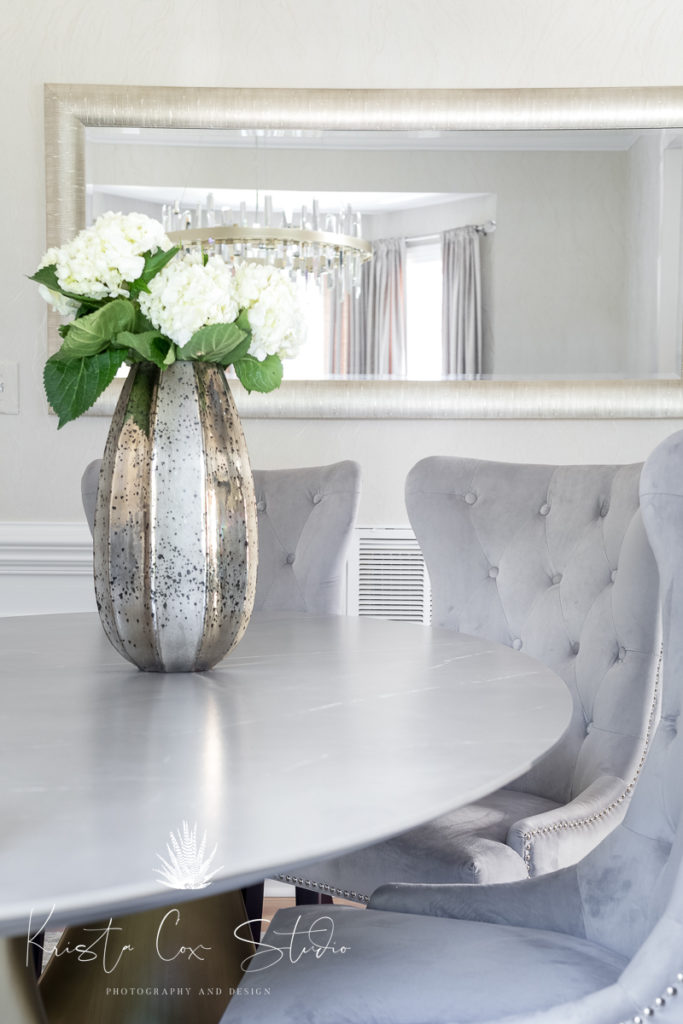Close up shot of vase of white flowers on dining table with mirror on wall behind. 