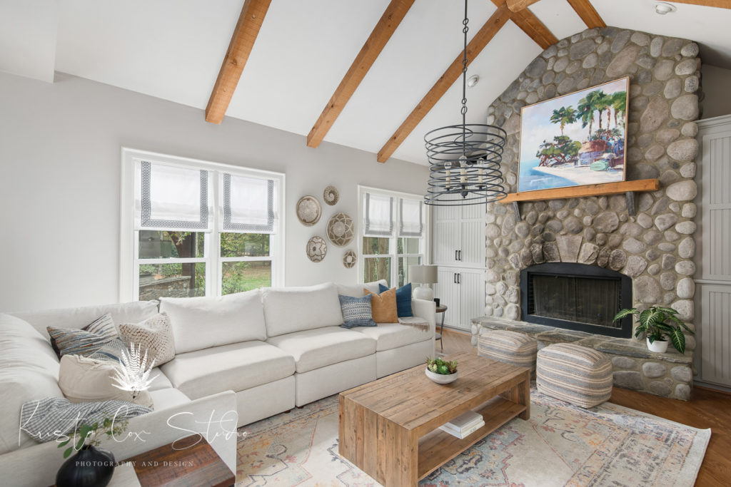 Angled shot of entire Livingroom, shows stone fireplace, vaulted ceilings with beams and large, white  L-shaped couch .